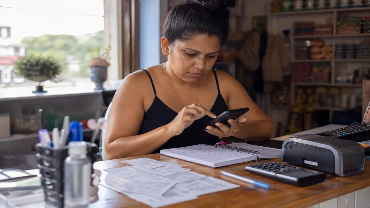 Microempreendedora utiliza celular para acessar portal gov.br, com comprovantes organizados em mesa de regularização de MEI.
